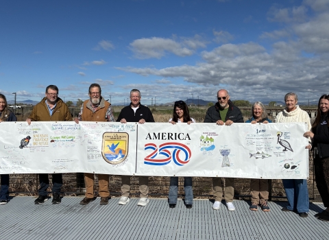 A group of people holds a banner with agency logo and that reads America 250 while standing on a deck with wetlands visible in the background. 
