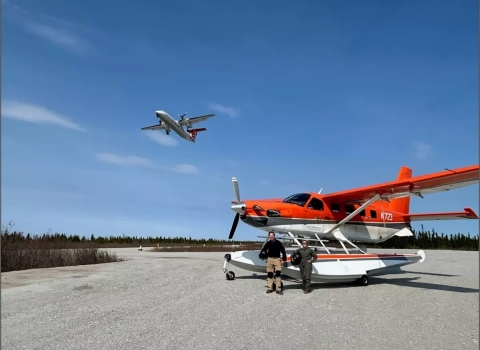 plane flying over two pilot biologists standing in front of a plane