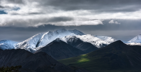 Snow-capped mountains loom over a green valley, all under stomy clouds